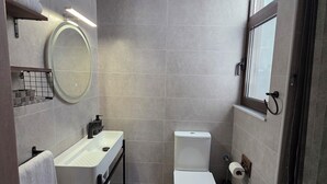 Un baño pequeño con lavamanos, espejo y retrete blancos. Hay un perchero sobre el lavamanos y una ventana a la derecha.