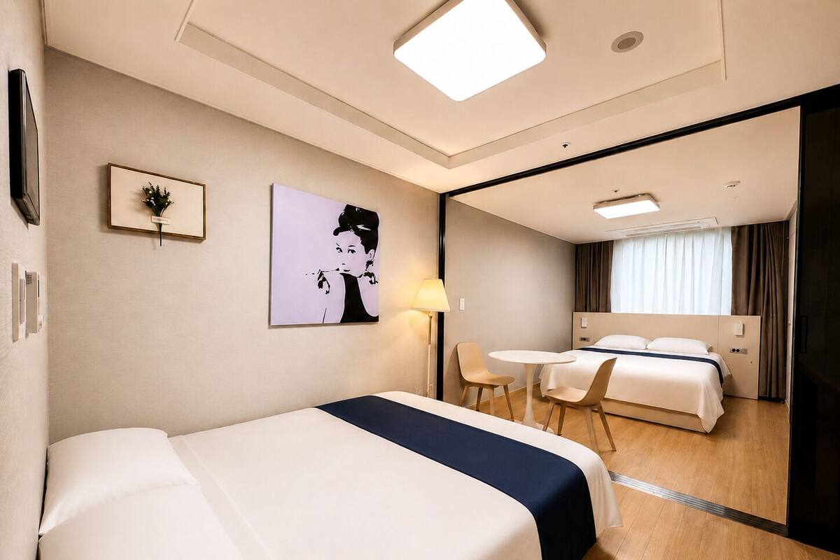 Habitación de hotel con cama, escritorio, dos sillas, lámpara, un cuadro y ventana con cortinas.