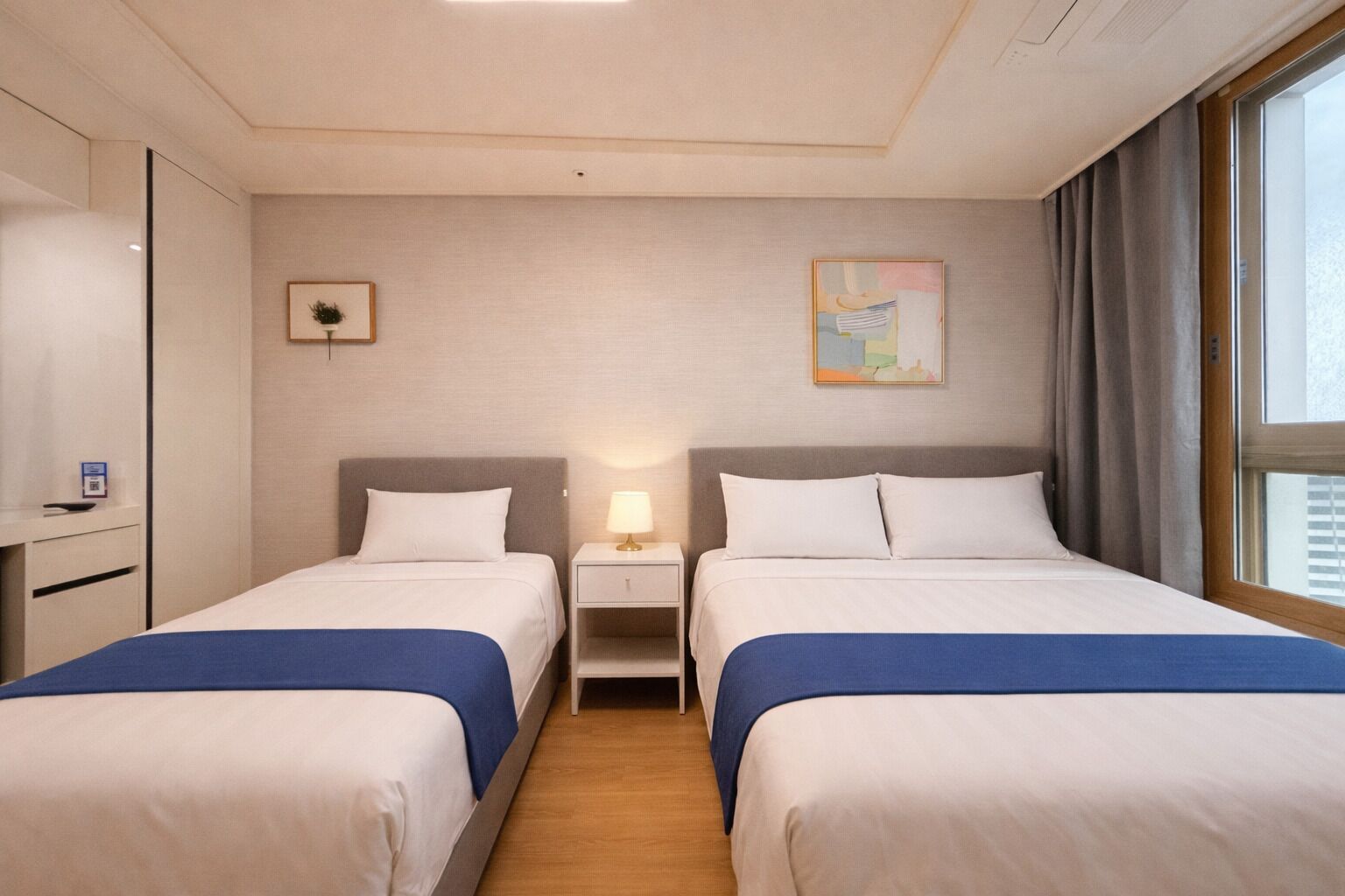 A hotel room with two beds, a desk, a lamp, and a painting on the wall.