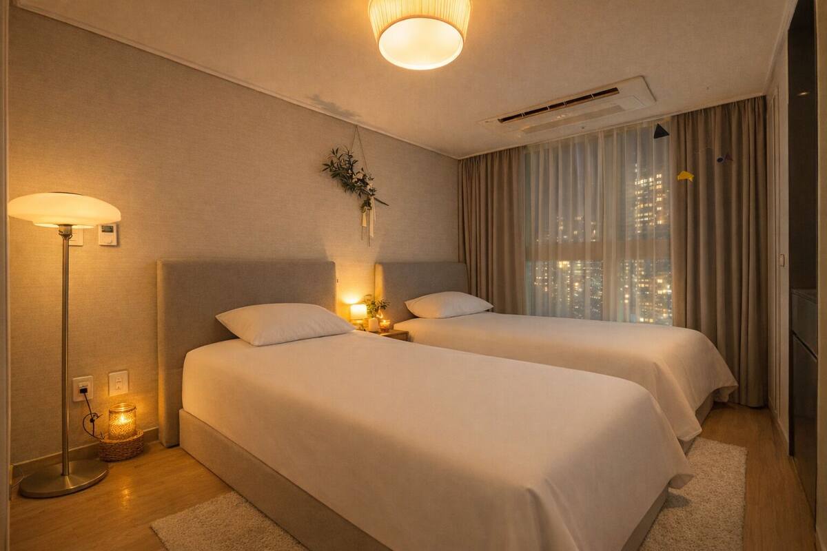 Habitación de hotel con dos camas, una mesita de noche con lámpara, una planta en la pared y vista nocturna de la ciudad.