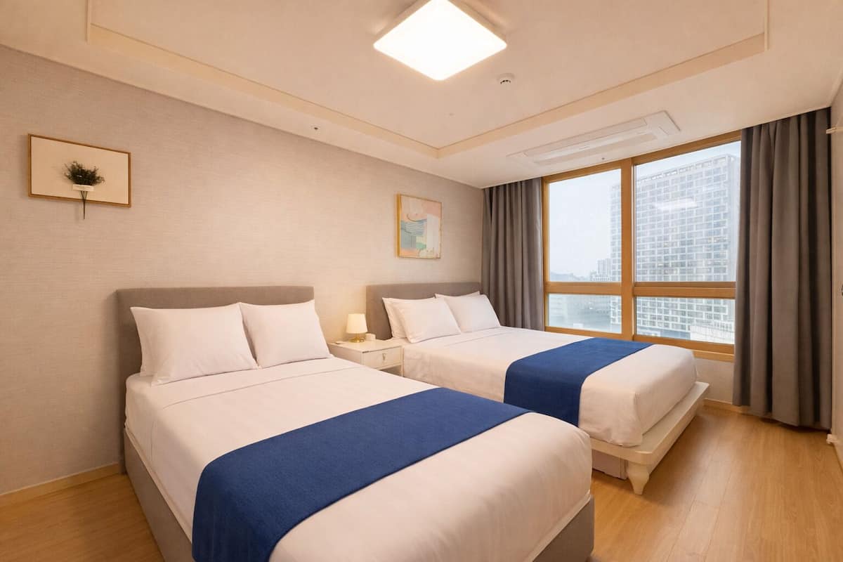 Habitación de hotel con dos camas, un ventanal grande con vistas a la ciudad, un cuadro en la pared y una lámpara en el techo.