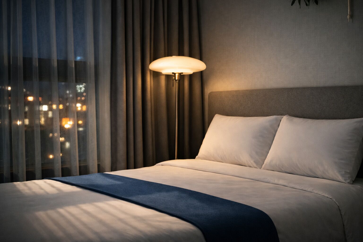 A hotel room with a bed, a nightstand, a lamp, and a view of the city at night.
