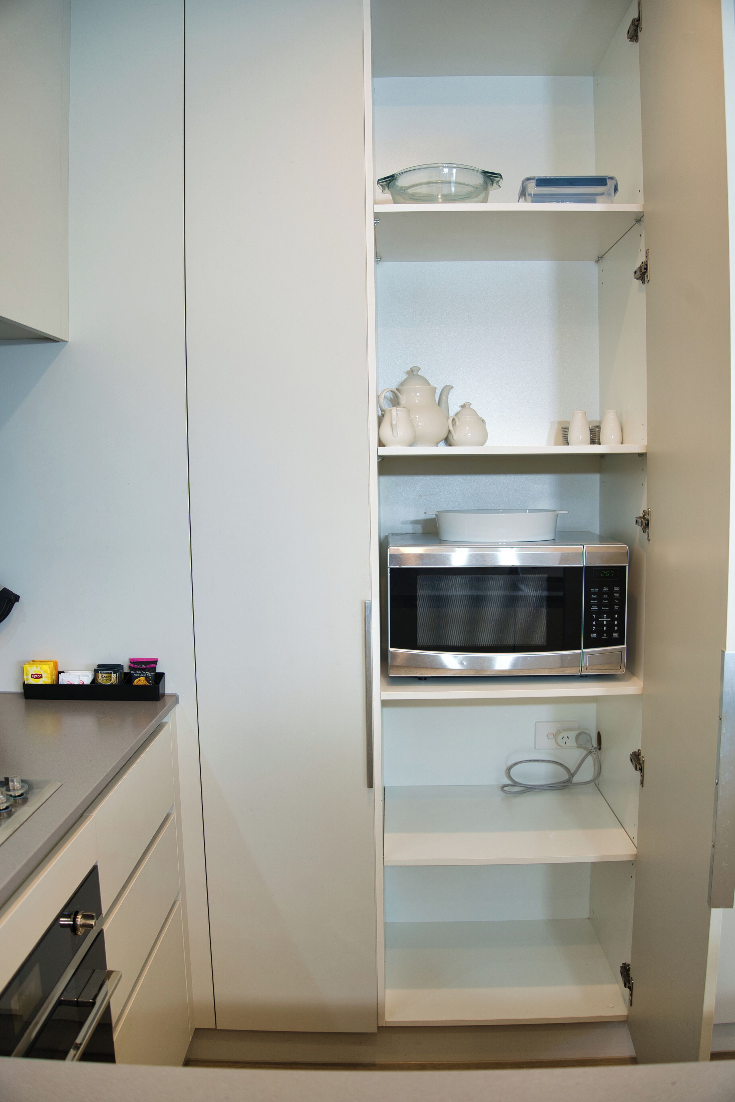 Una cocina con un microondas en el estante central, un bol de vidrio en el estante superior y algunos utensilios de cocina en los estantes inferiores.