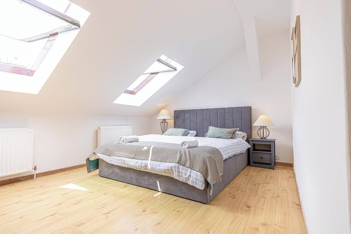 Ein Schlafzimmer mit Oberlicht, einem Bett mit grauem Kopfteil, zwei Nachttischen mit Lampen und einer Heizung.
