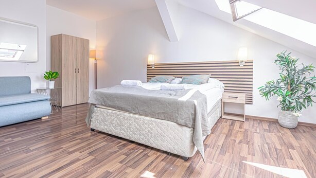 Une chambre moderne avec un plafond de style industriel, un lit doté d’une tête de lit en bois, une table de chevet avec une lampe, une petite plante et un canapé bleu.