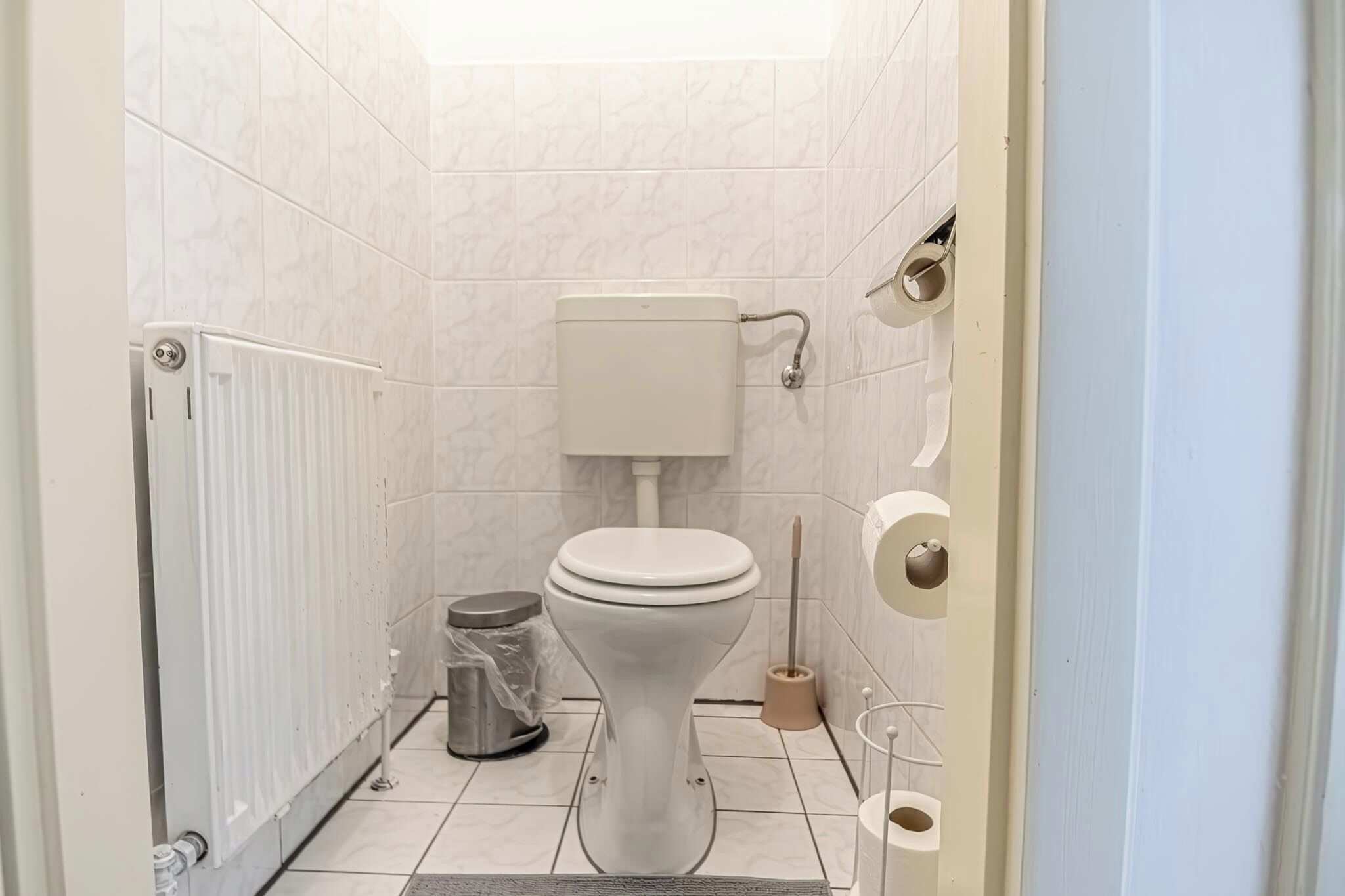 A small, modern bathroom with a toilet, radiator, trash bin, and toilet brush.