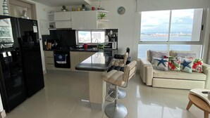 A modern kitchen with a central island, a dining area with bar stools, a sofa with decorative pillows, and a large window with a city view.