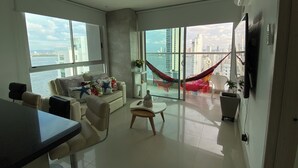 A modern living room with a sofa, armchairs, a coffee table, and a TV. Large windows offer a view of a cityscape and a body of water.