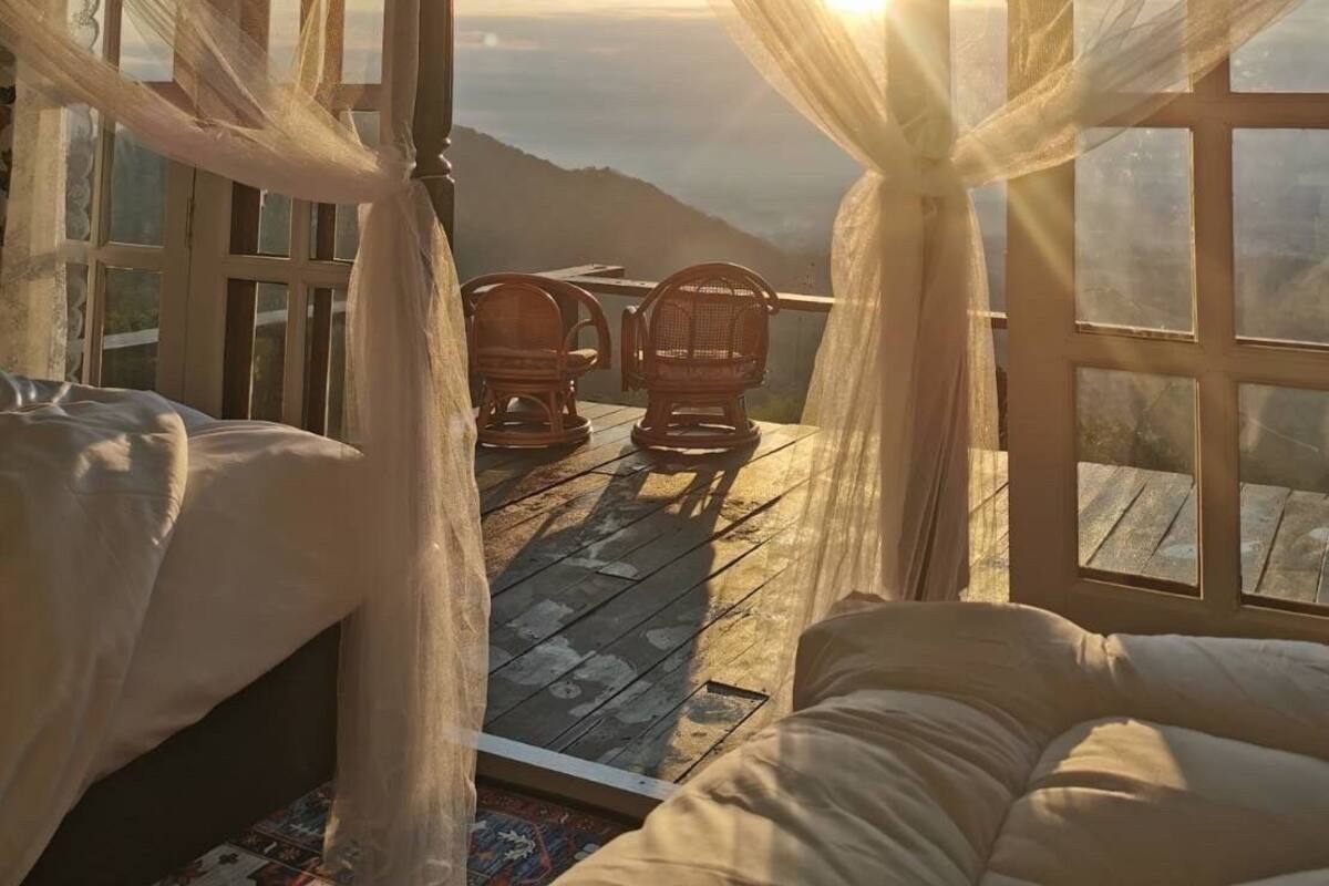 Une chambre avec un lit, deux fauteuils en osier et une vue sur les montagnes, visible à travers de grandes fenêtres.