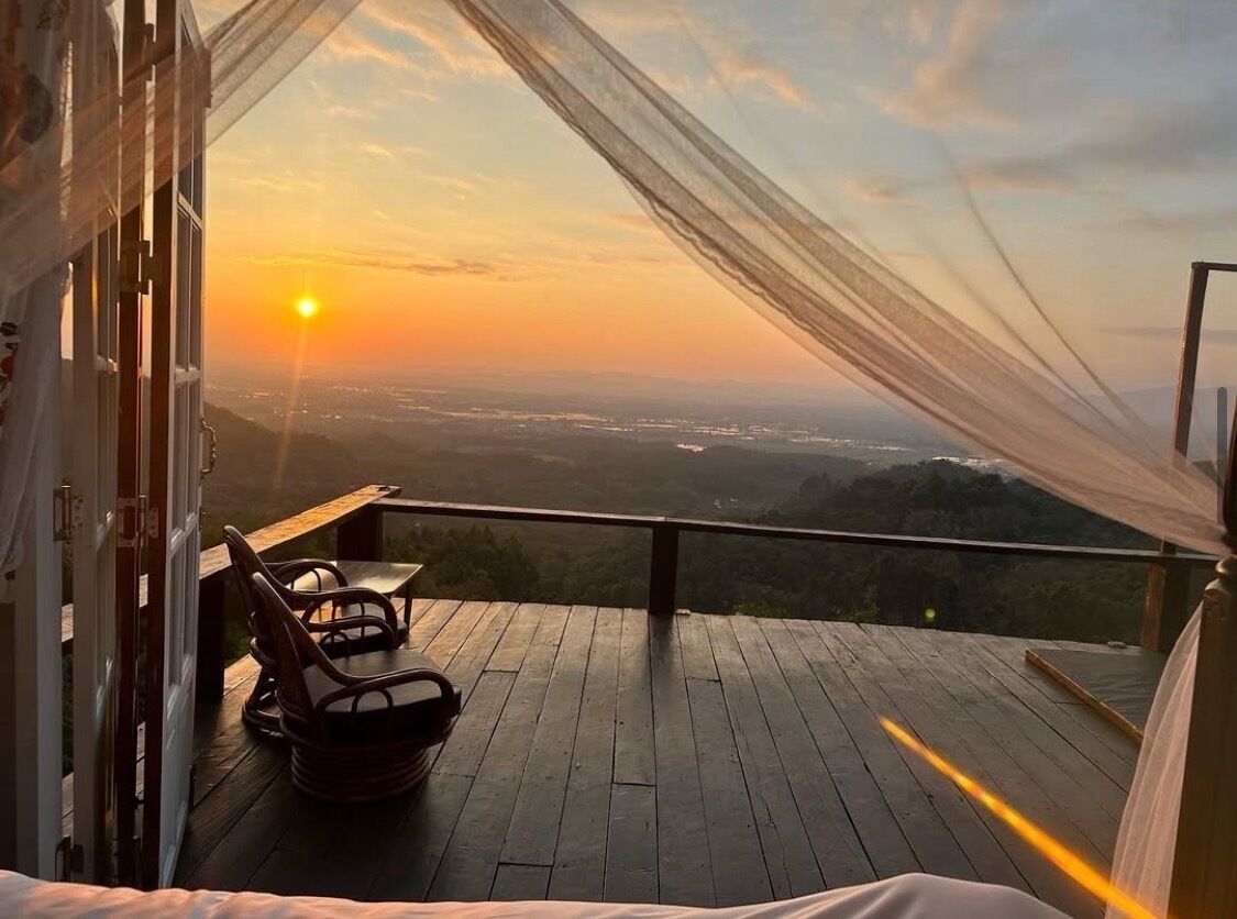 A balcony with a wooden floor and two wicker chairs, overlooking a scenic view of a sunset over a valley.
