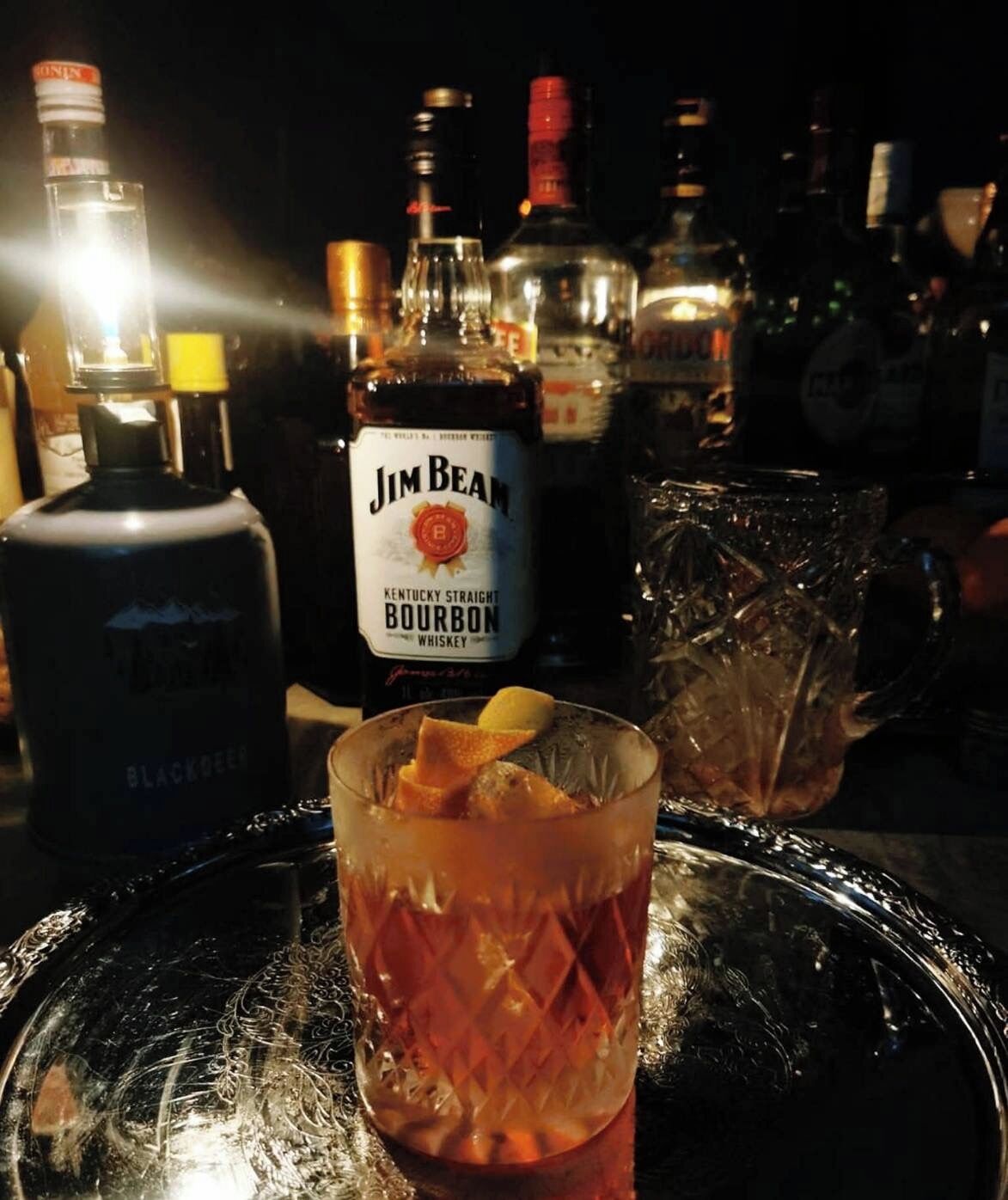 A bar setting with a bottle of Jim Beam Kentucky Straight Bourbon Whiskey and a glass with ice and a lemon wedge.