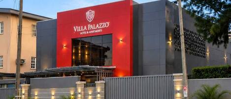 Un hotel moderno con una fachada roja destacada, una entrada protegida por una reja y un área exterior ajardinada.