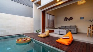 Een modern buitenzwembad met een houten terras, een grijze bank en een rieten mand met lekkernijen.