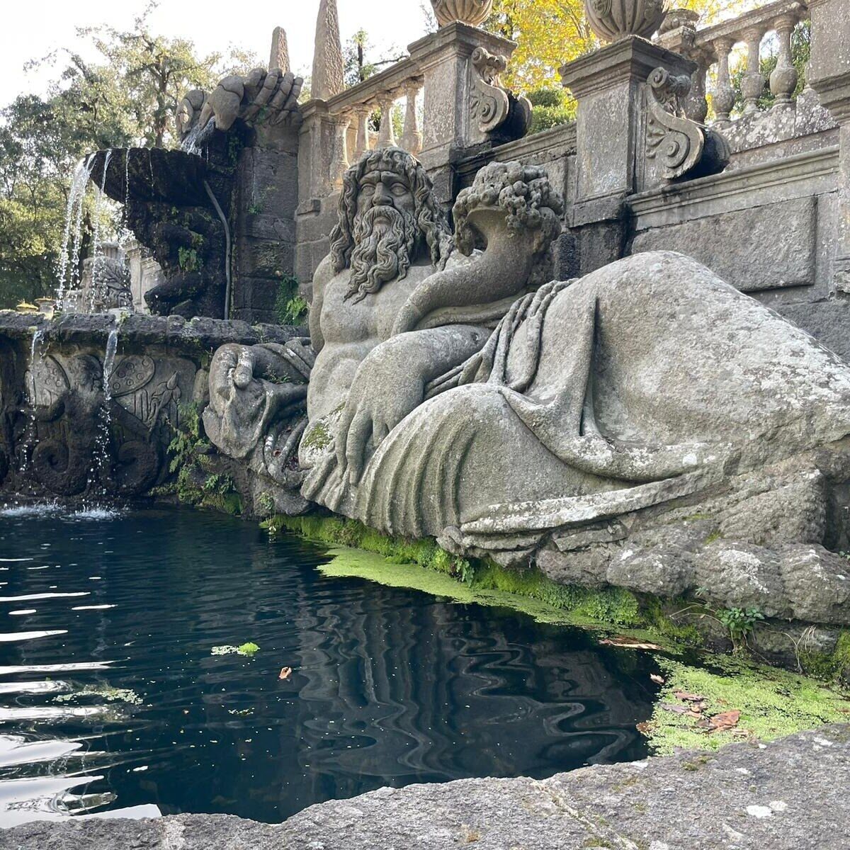 Eine Steinskulptur eines liegenden menschlichen Körpers neben einem Wasserspiel, mit einer Brücke und aufwendigen Brüstungen im Hintergrund.