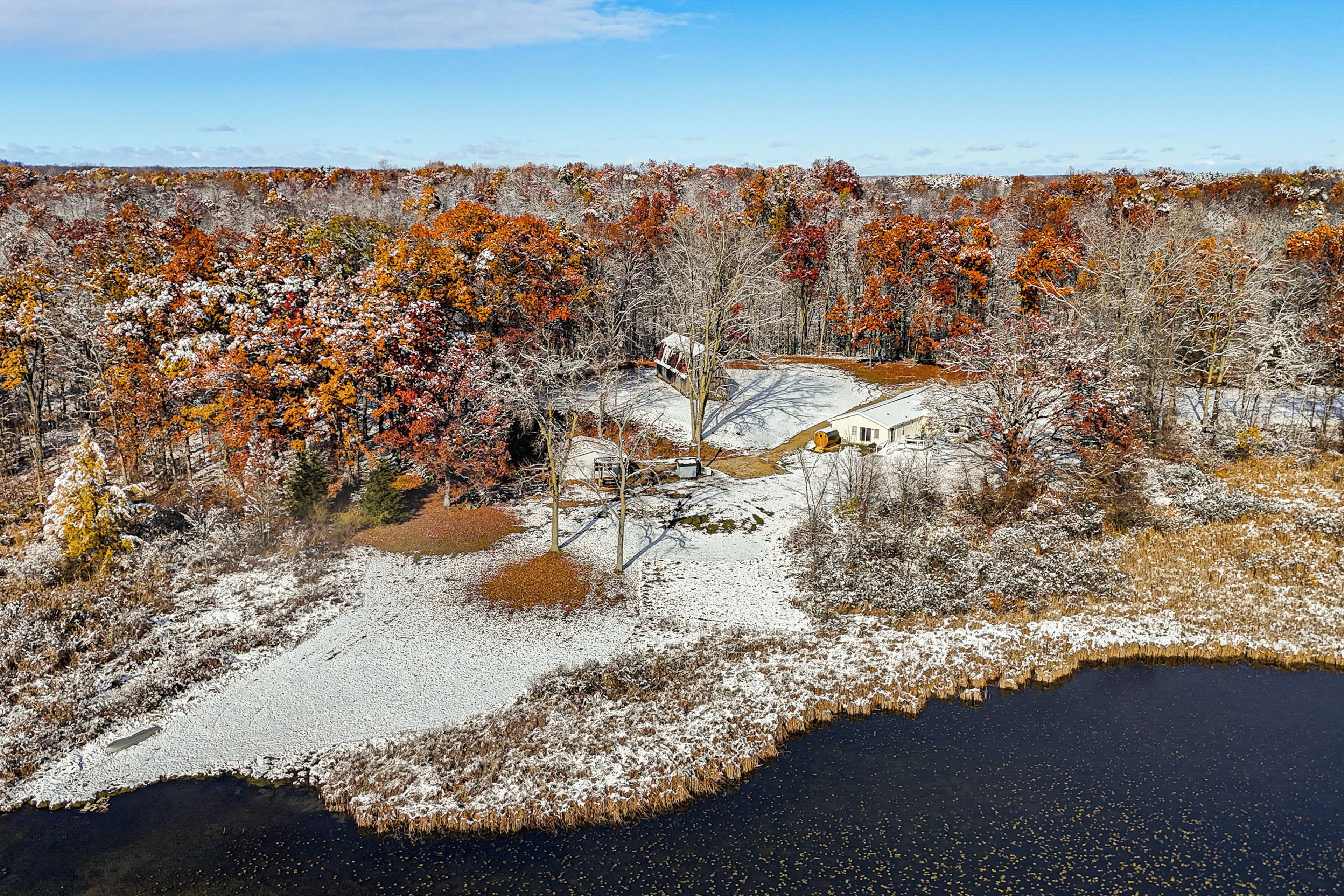 A winter landscape with a snow-covered pond, trees with autumn foliage, and a house in the background.