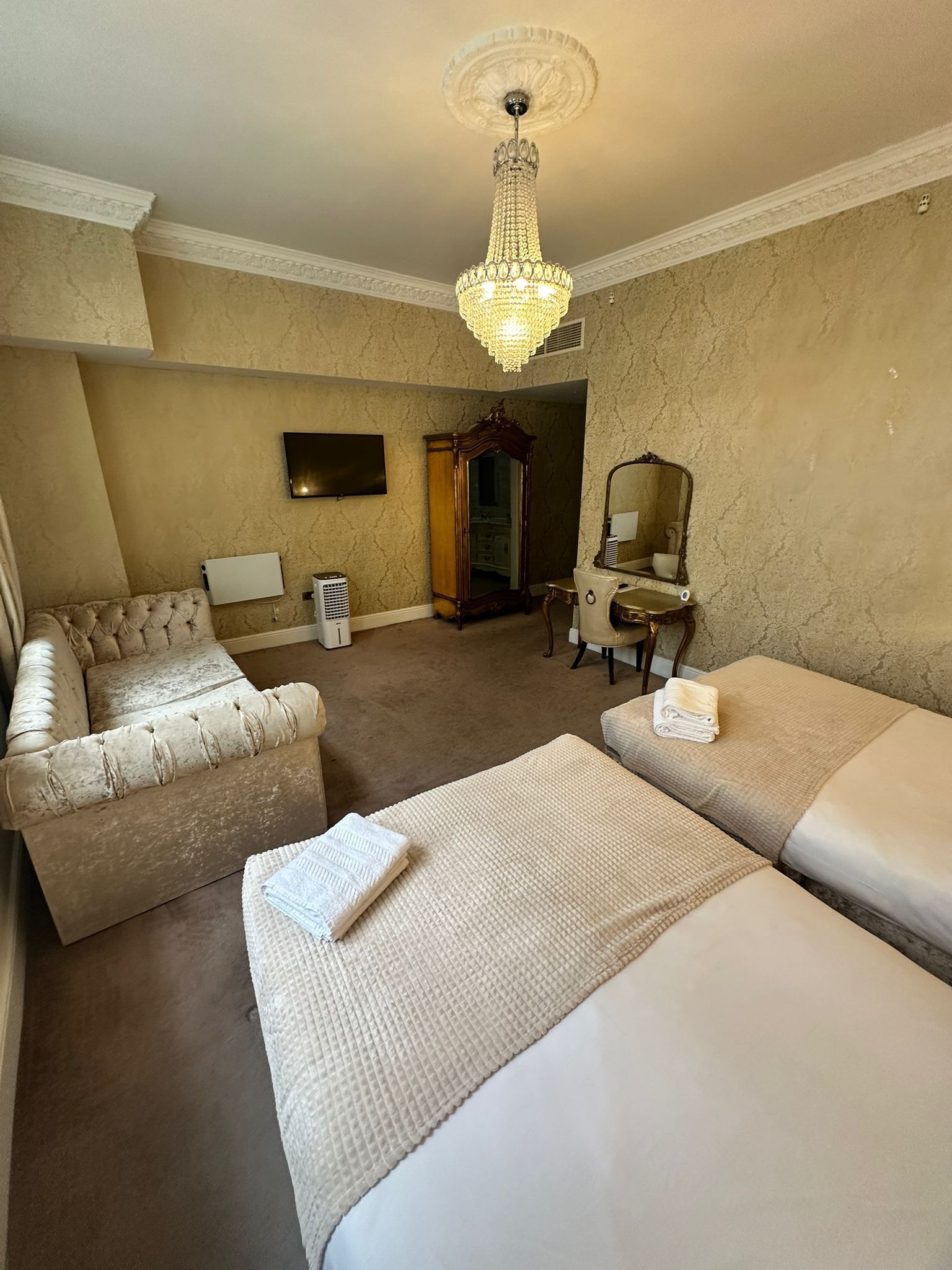 A hotel room with a large bed, a sofa, a desk, a mirror, a chandelier, and a television.