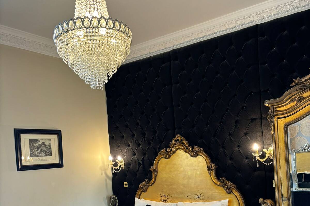 Un dormitorio lujoso con una cama grande, una araña, un cabecero tapizado oscuro, un espejo con marco dorado y un cuadro en la pared.