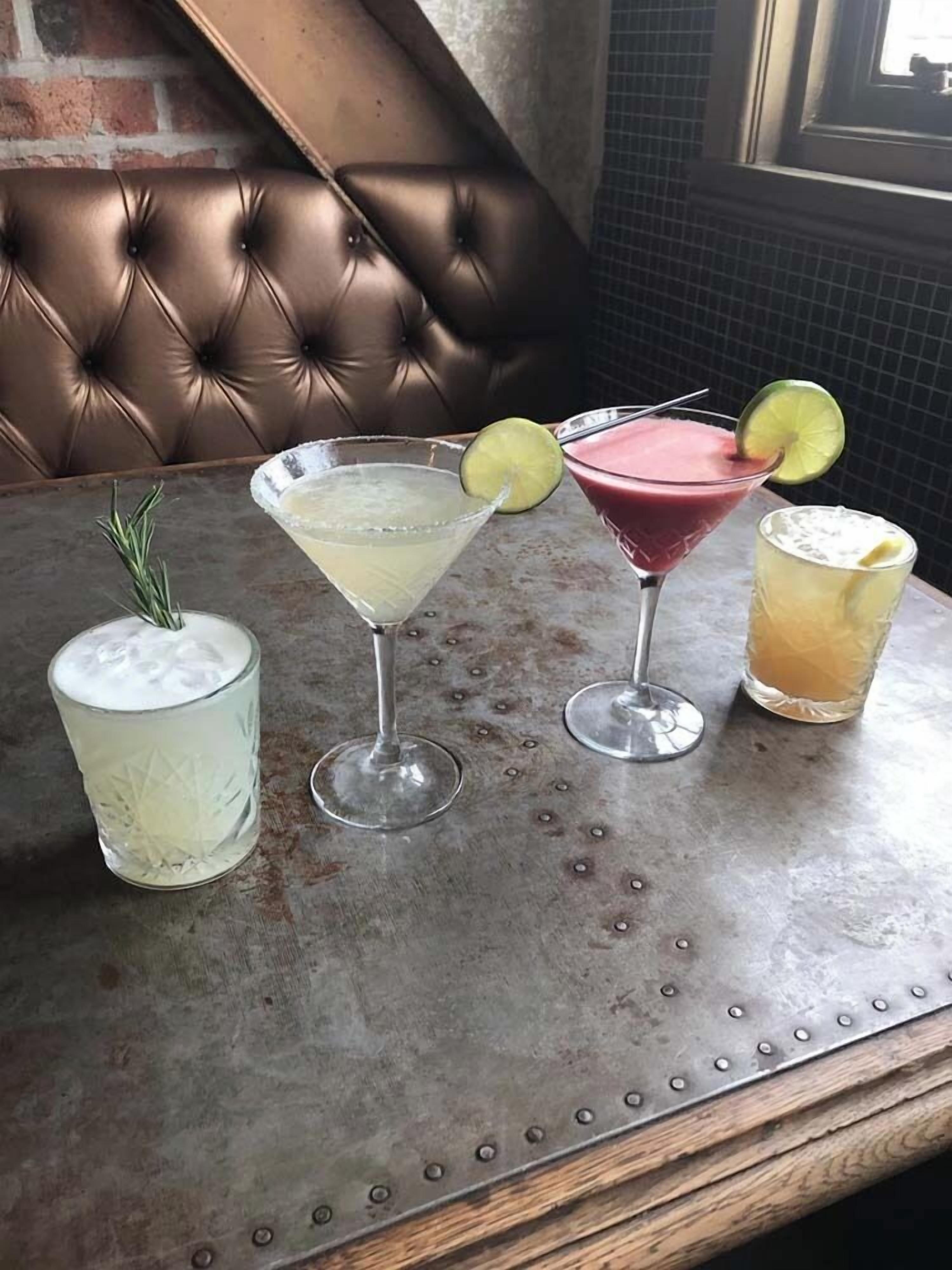 Een tafel met vier drankjes, een glas met een rozemarijn takje, en een bruine leren bank op de achtergrond.