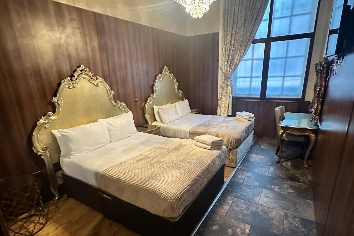 Habitación de hotel con dos camas, un candelabro, un ventanal grande con cortinas y una pequeña mesa con una silla.