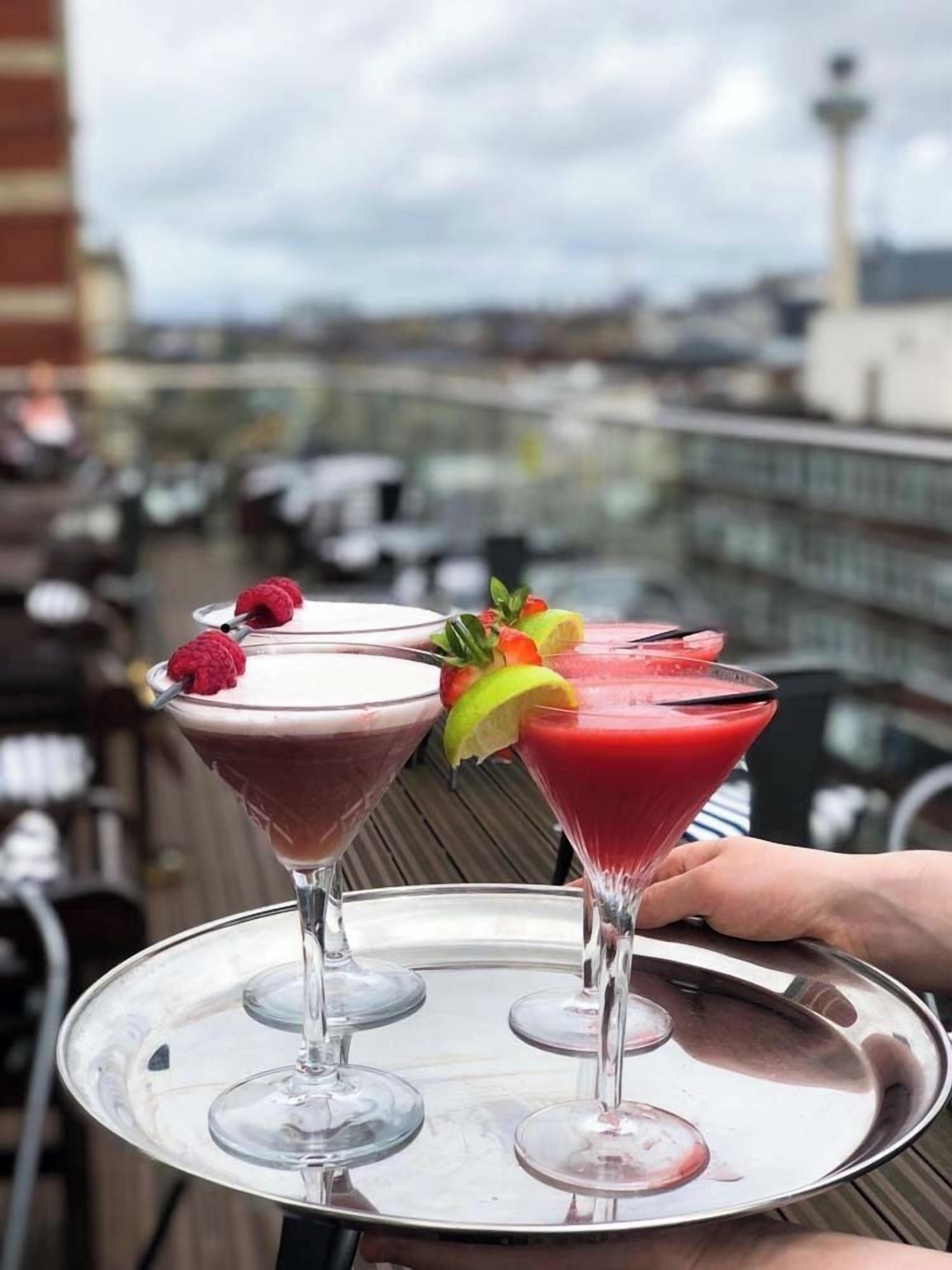 Ein Tablett mit drei Martini-Gläsern, die unterschiedliche Getränke enthalten, mit Früchten garniert und von einer Person gehalten.