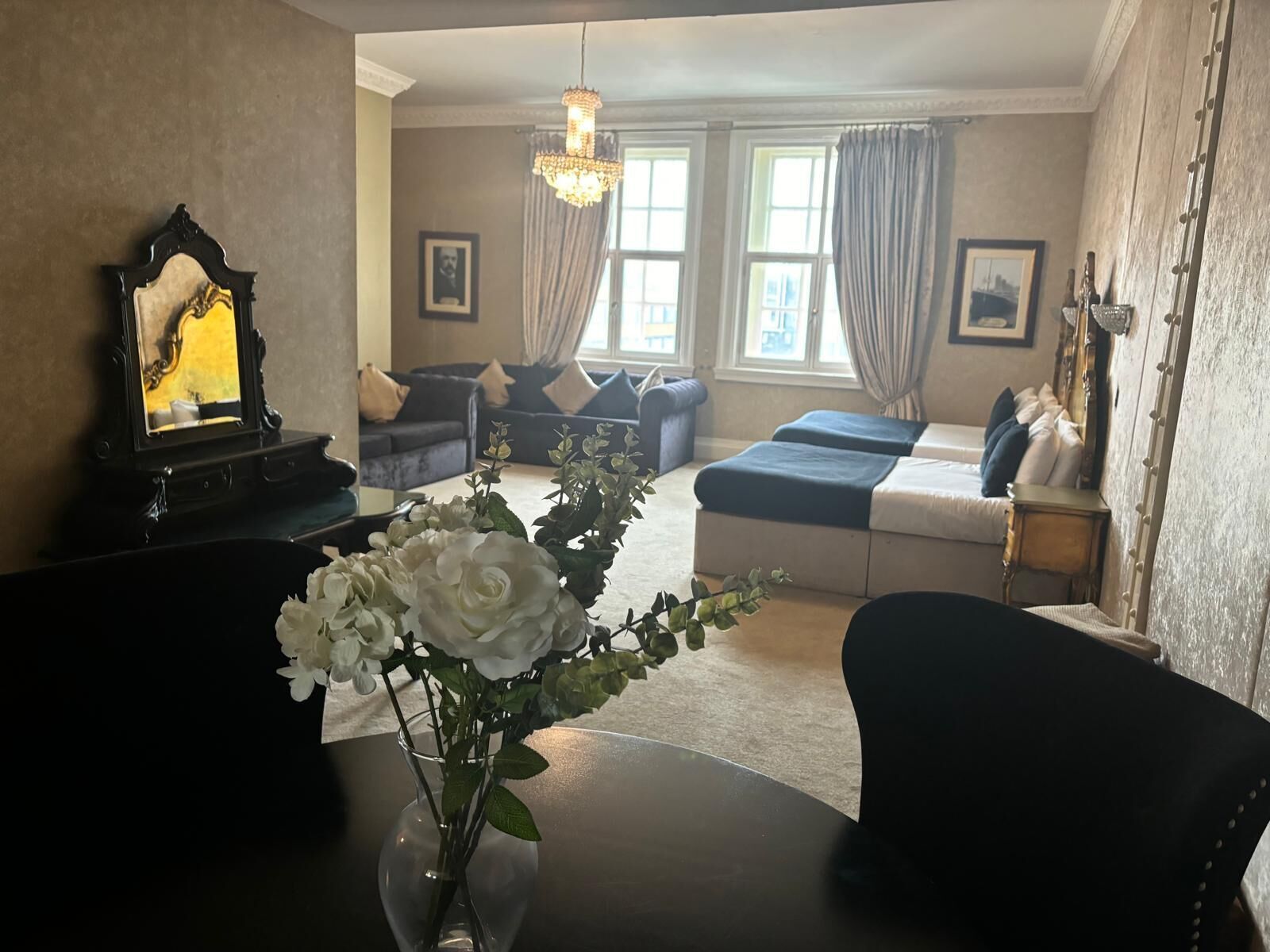 Een hotelkamer met een bed, een bank, een stoel, een tafel met een bloemenvaas, een spiegel en een kroonluchter.