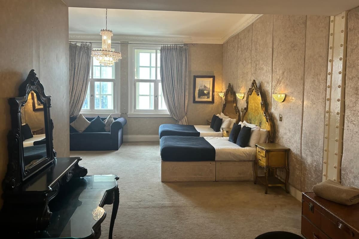 Una habitación de hotel con un piano, dos camas, una araña y un gran ventanal con cortinas.