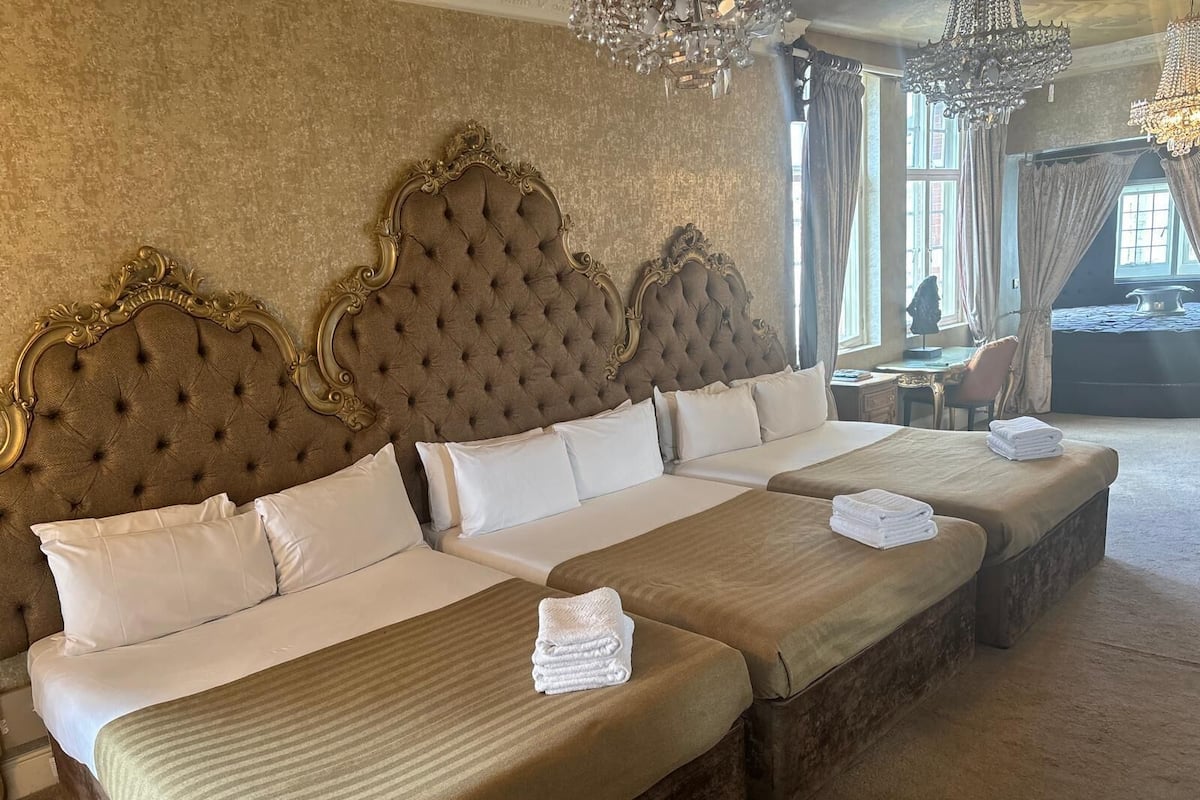 Una habitación de hotel con tres camas grandes, cada una con un cabecero capitonado y ropa de cama blanca. Hay dos candelabros, un ventanal con cortinas y vistas al exterior.