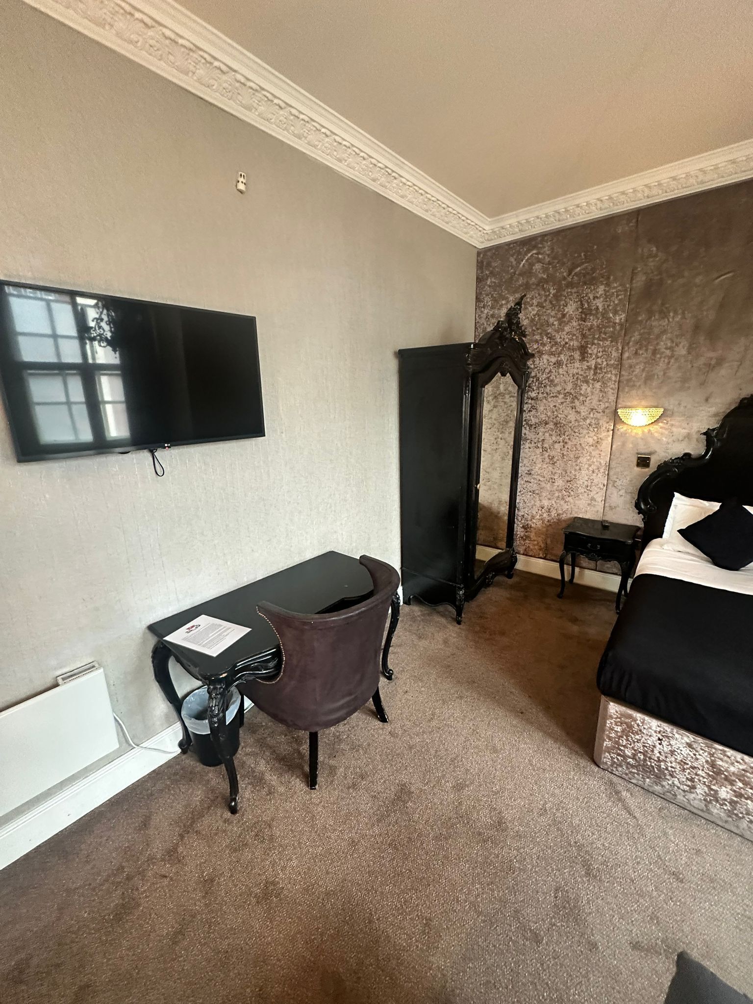 A hotel room with a flat-screen TV mounted on the wall, a desk with a chair, a bed, and a nightstand.