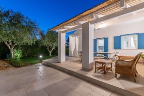 Un patio techado al aire libre con una mesa y sillas, una estructura de pérgola y vistas del jardín y los árboles circundantes.