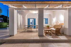 Un patio techado al aire libre con una mesa y sillas, una silla de mimbre y una pequeña área de cocina con un lavamanos y elementos decorativos.