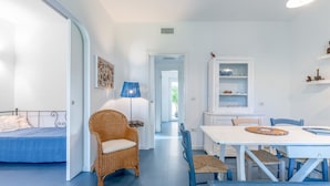 Une pièce comprenant un lit, une chaise en osier, une table avec des chaises, une lampe et un meuble avec des étagères.