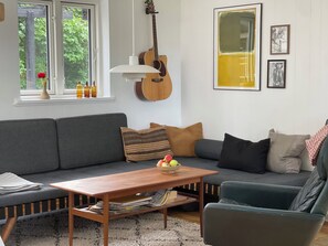 A living room with a grey sofa, a wooden coffee table with books, a guitar, and framed pictures on the wall. - Lovely year-round home with sea view (Føllenslev)