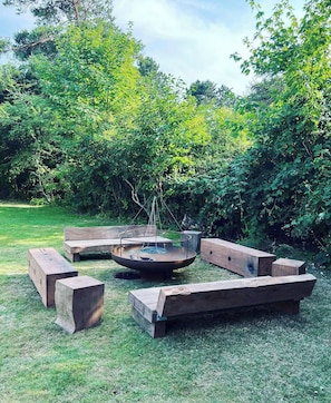 A circular fire pit with benches around it, set in a grassy outdoor area with dense foliage in the background.