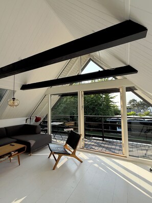 A modern loft bedroom with a sloped ceiling, large windows, a sofa, a chair, and a table.