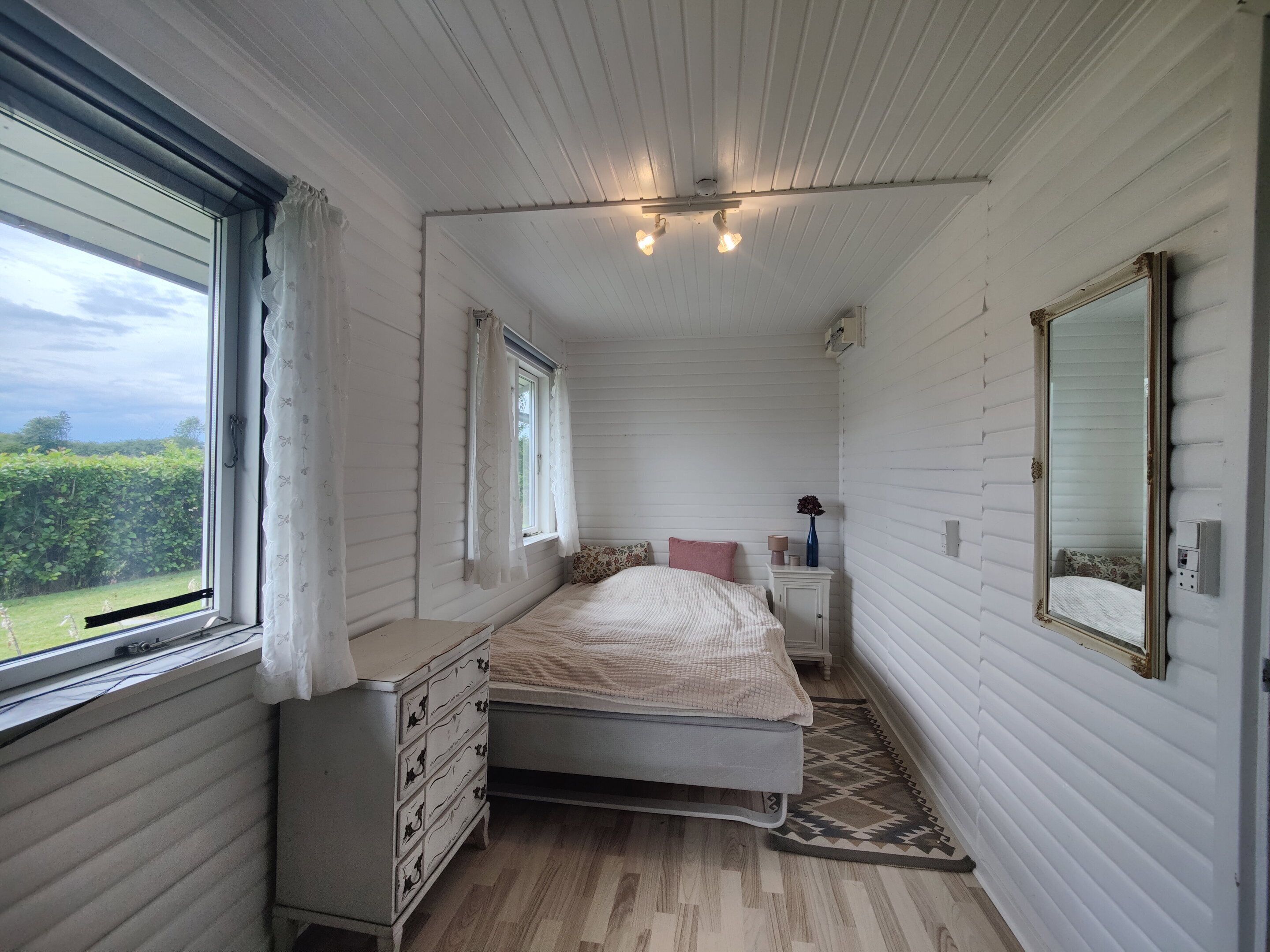 A bedroom with a bed, dresser, bedside table, mirror, and a window with curtains.