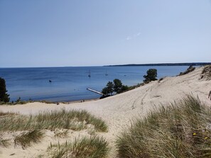 En strand med sanddyner, gles vegetation och stilla vatten.
