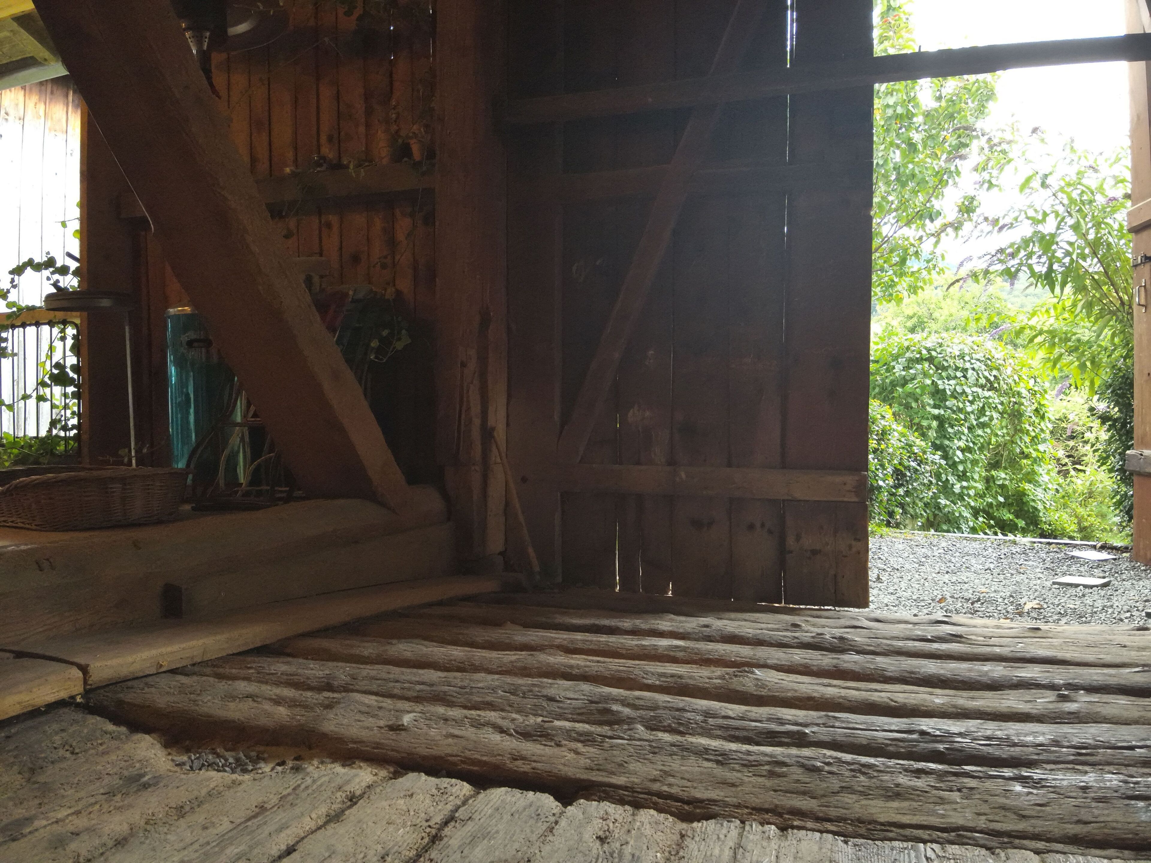 A rustic wooden barn interior with a wooden floor, a large open door leading outside, and a metal water tank visible through the door.