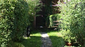 A pathway leading to a building with a covered entrance, surrounded by lush greenery and flowering plants. - Vacation apartment with terrace and mountain view (Frasdorf)