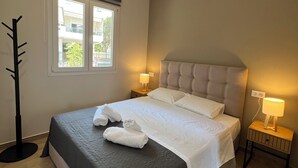 Una cama bien hecha con sábanas blancas y dos toallas, una lámpara de noche y una ventana con vistas a edificios y árboles.