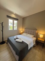 A neatly made bed with white linens and two towels, a bedside lamp, and a window with a view of buildings and trees.