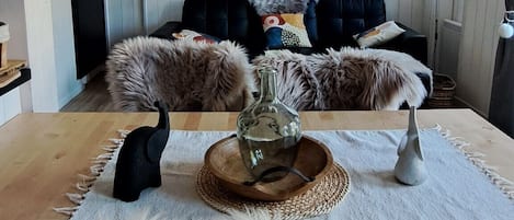 A rustic interior with a high ceiling, wooden beams, a large dining table with a white table runner, fluffy white rugs, and a fireplace.
