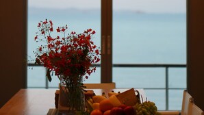 Une table à manger ornée d’un vase de fleurs rouges, d’un saladier de fruits et offrant une vue sur la mer grâce à de grandes fenêtres.