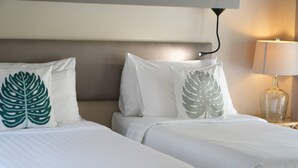 Habitación de hotel con dos camas, cada una con ropa de cama blanca y almohadas con estampado de hojas. Una lámpara de noche está a la derecha.