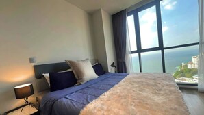 Een slaapkamer met een groot bed, een nachtkastje, een lamp en uitzicht op de zee door een groot raam.