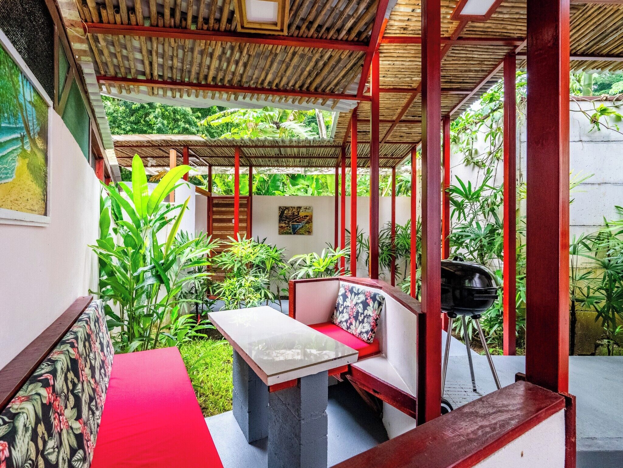 A covered outdoor seating area with a table and chairs, surrounded by lush greenery.