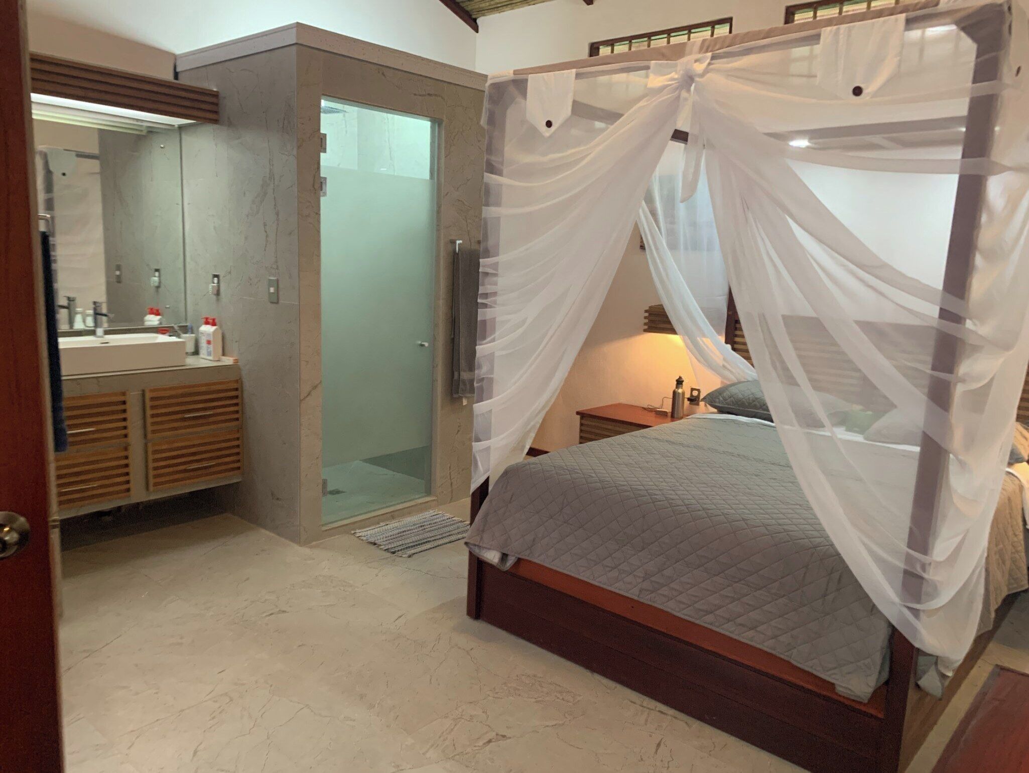 A bedroom with a bed, a nightstand, a lamp, a mirror, a shower, and a vanity with a sink.