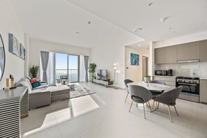 A modern living room with a sofa, dining table, and chairs, a kitchen with appliances, and large windows offering a view. - Luxury City Stay Near Mall and Pool (Dubai)