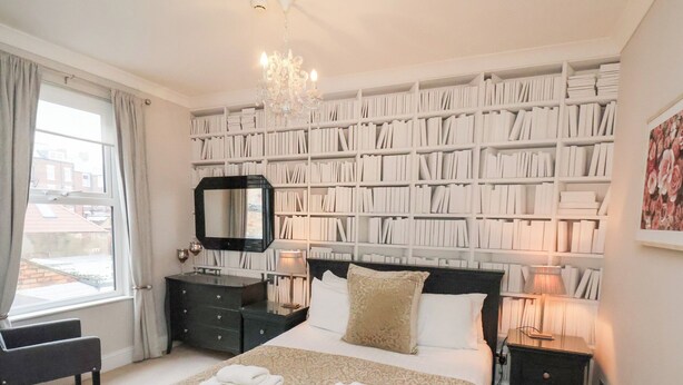 A bedroom with a large bookshelf, a bed with white linens, a dresser, and a chandelier. - All Seasons (Filey)