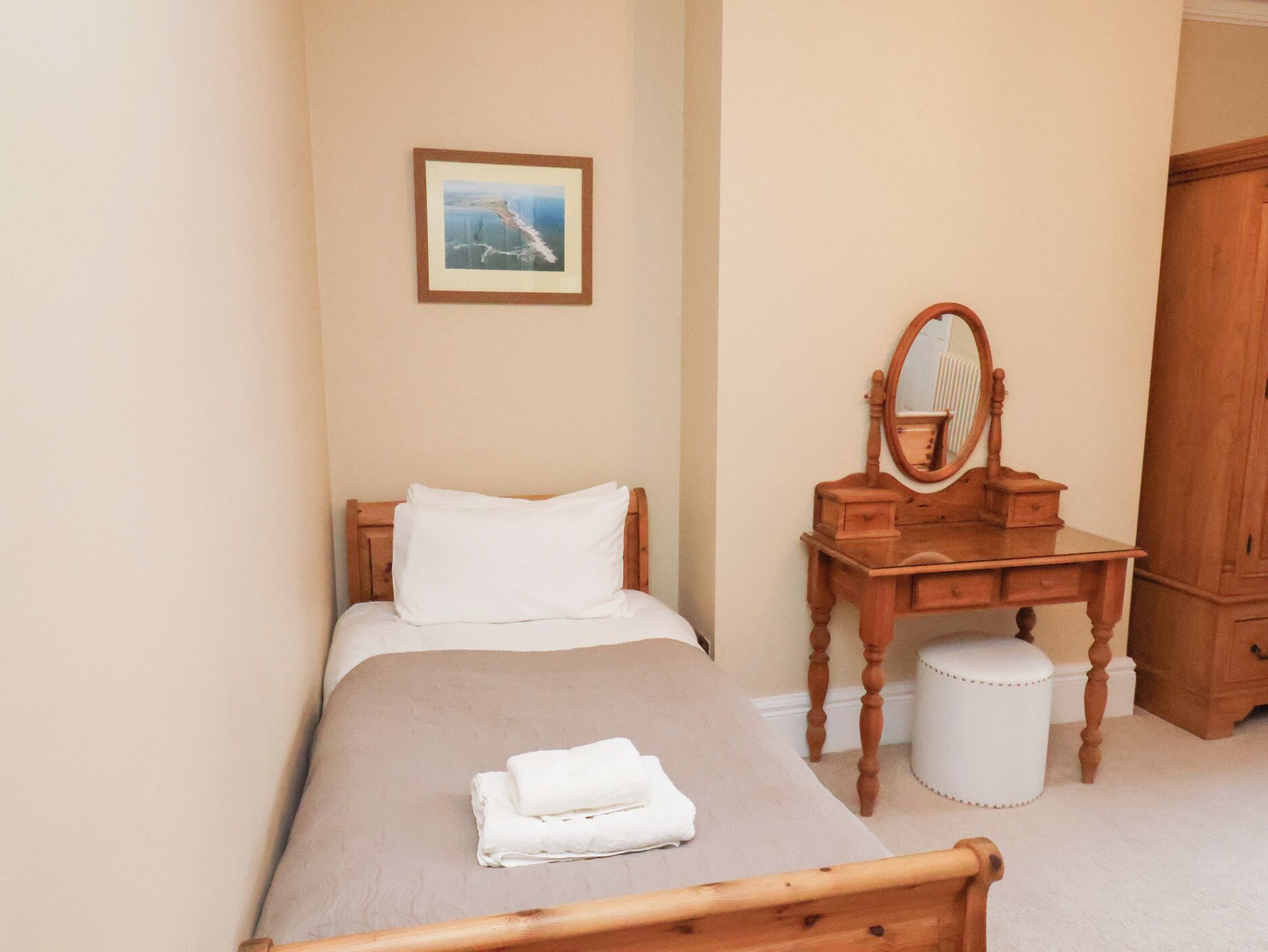 A bedroom with a bed, a wooden dresser with a mirror, a small round stool, and a framed picture on the wall.
