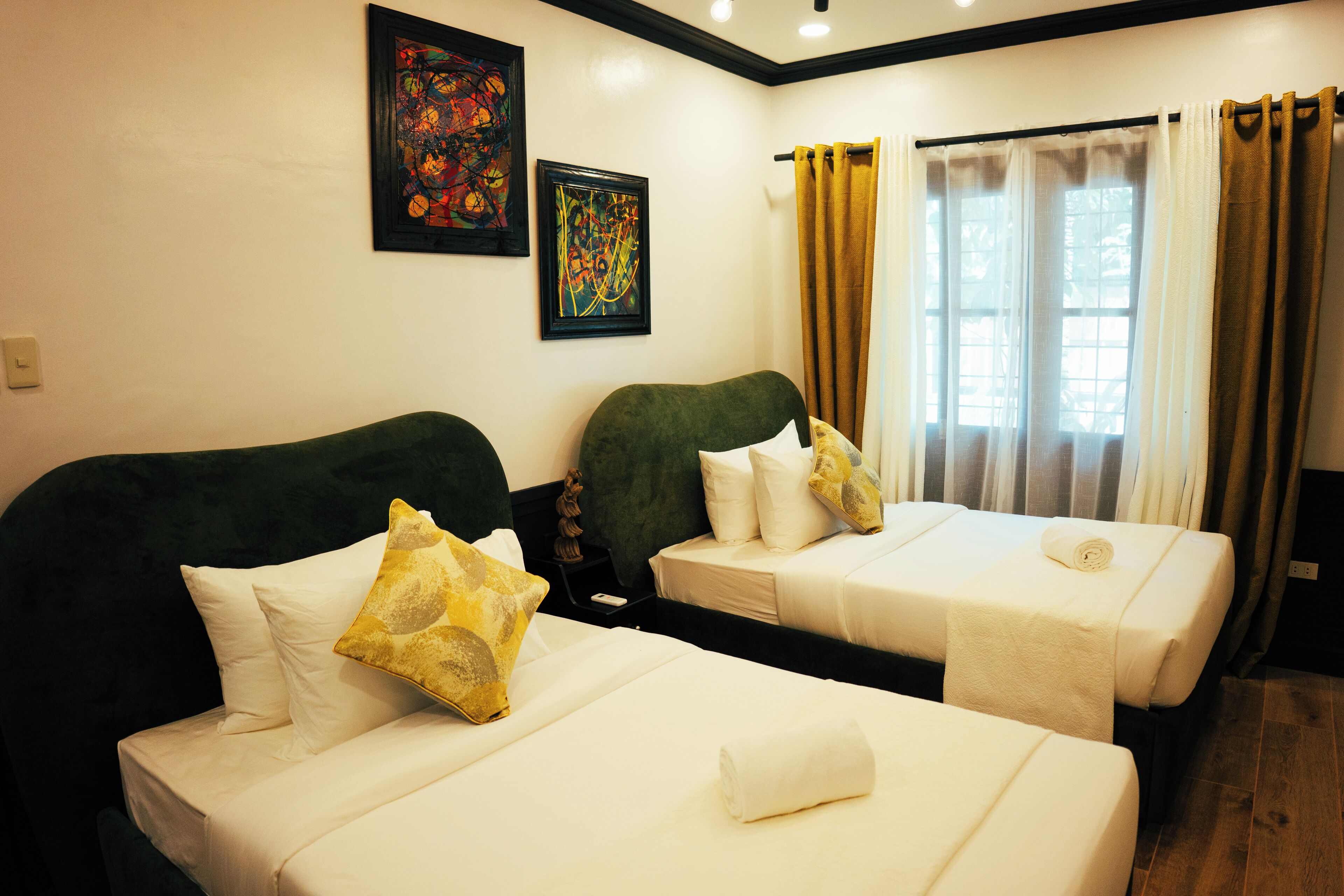 Een hotelkamer met twee bedden, elk voorzien van witte lakens en decoratieve kussens. Twee ingelijste abstracte schilderijen hangen aan de muur, en er is een raam met gordijnen.
